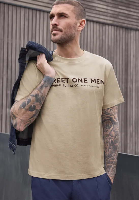 Mann mit beige T-Shirt 'STREET ONE MEN' und Denimjacke über der Schulter vor Holzwand