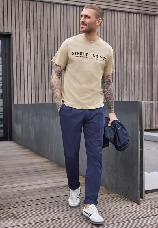 Ein tätowierter Mann in beige T-Shirt und blauen Hosen läuft mit Jacke in der Hand.