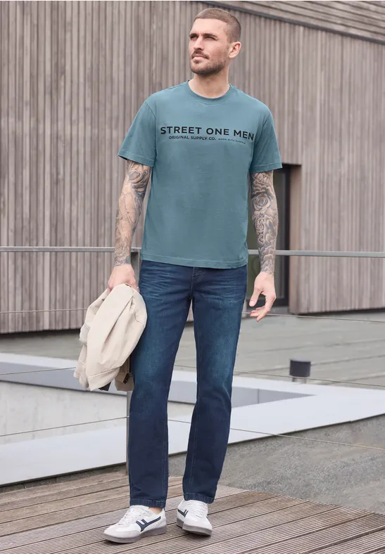 Mann in taubenblauem T-Shirt mit Aufschrift, Jeans und Sneakern steht vor Holzwand und hält Jacke.