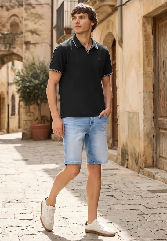 Ein Mann in schwarzem Poloshirt und Jeansshorts spaziert durch eine gepflasterte Gasse einer alten Stadt.