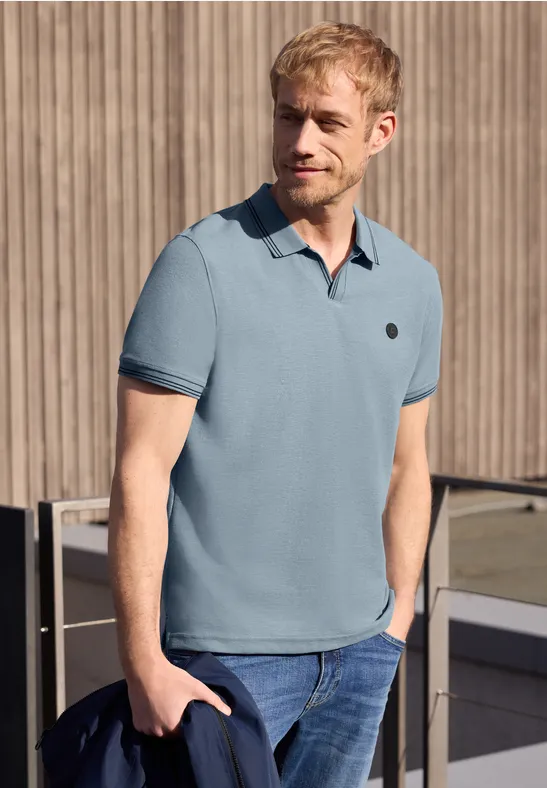 Ein Mann trägt ein hellblaues Poloshirt und Jeans, steht vor einer Holzverkleidung.