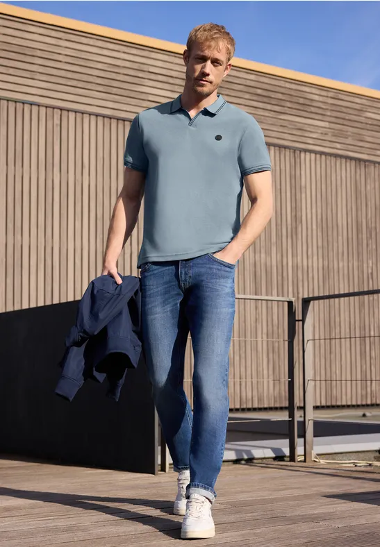 Ein Mann in blauem Poloshirt, Jeans und Sneakern steht auf einer Holzterrasse und hält eine Jacke in der Hand.
