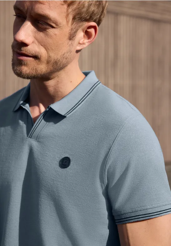 Ein Mann mit leichtem Bart trägt ein hellblaues Poloshirt vor einem holzverkleideten Hintergrund.