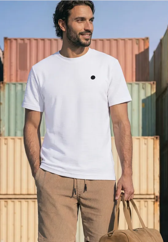 Ein Mann in weißem T-Shirt und beigen Hosen posiert vor bunten Containern mit Tasche.