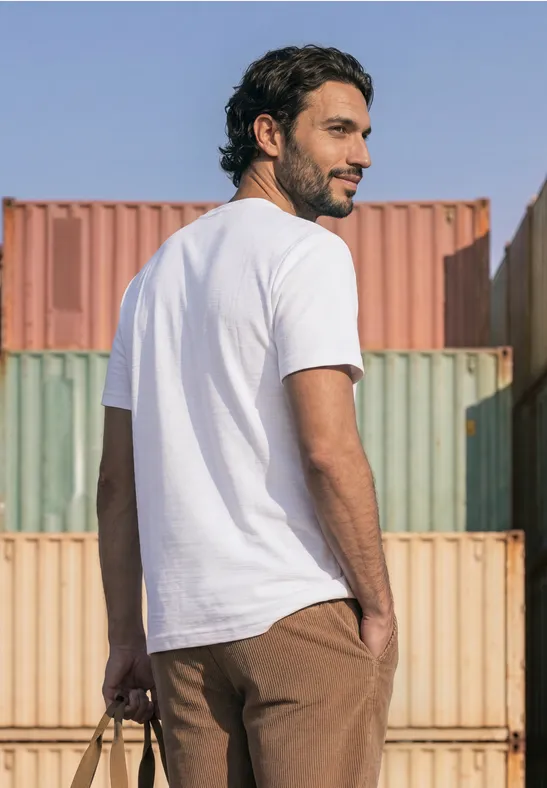 Ein Mann in weißem T-Shirt und brauner Hose steht vor bunten Containern, Blick zurückgeworfen.