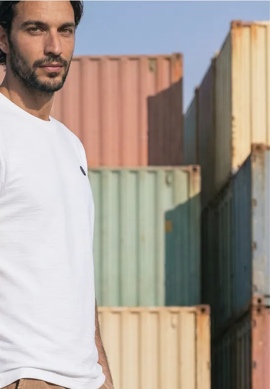 Ein Mann in weißem T-Shirt steht vor bunten Schiffscontainern.