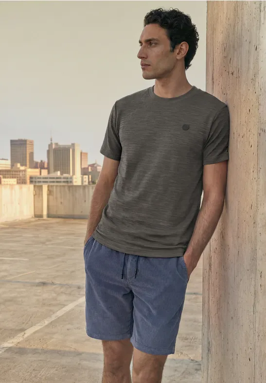 Ein Mann lehnt an einer Betonstütze auf einem Dach, trägt T-Shirt und Shorts.