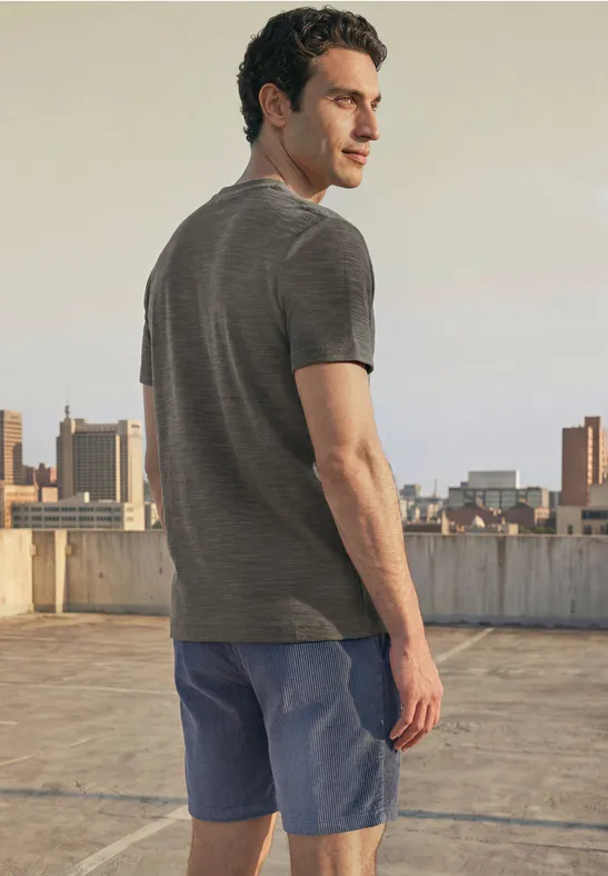 Ein Mann in grauem T-Shirt und blauen Shorts steht auf einem Dach mit Stadtpanorama und Blick über die Schulter.