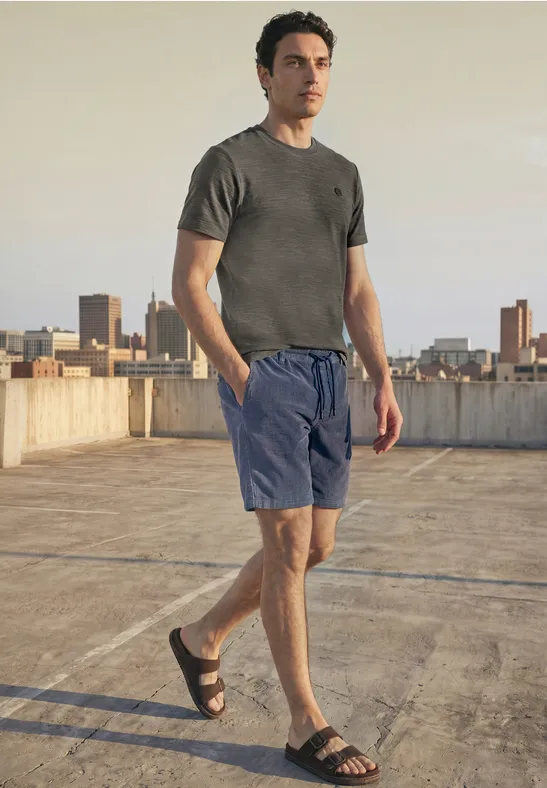 Ein Mann steht auf einem Parkdach, trägt graues T-Shirt, blaue Shorts und Sandalen, Skyline im Hintergrund.
