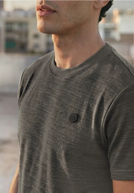 Ein Mann trägt ein graues T-Shirt mit kleinem Brustlogo, unscharfer urbaner Hintergrund.