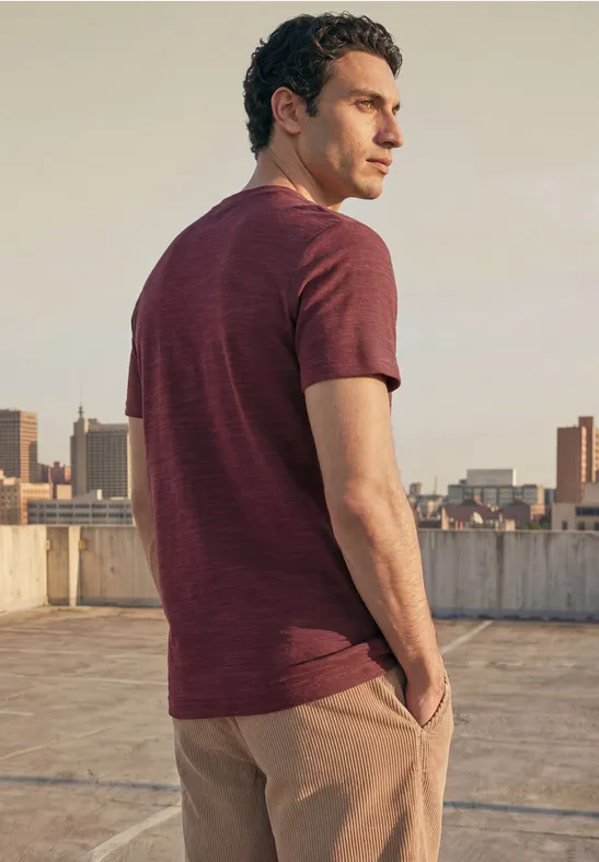 Ein Mann steht auf einem Dach, blickt über die Stadt und trägt ein rotbraunes T-Shirt mit beigefarbener Hose.