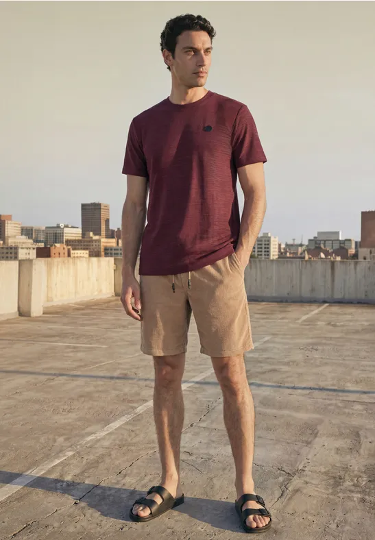 Ein Mann auf einem Parkdeck mit Stadtblick trägt ein bordeauxfarbenes T-Shirt, Shorts und Sandalen.