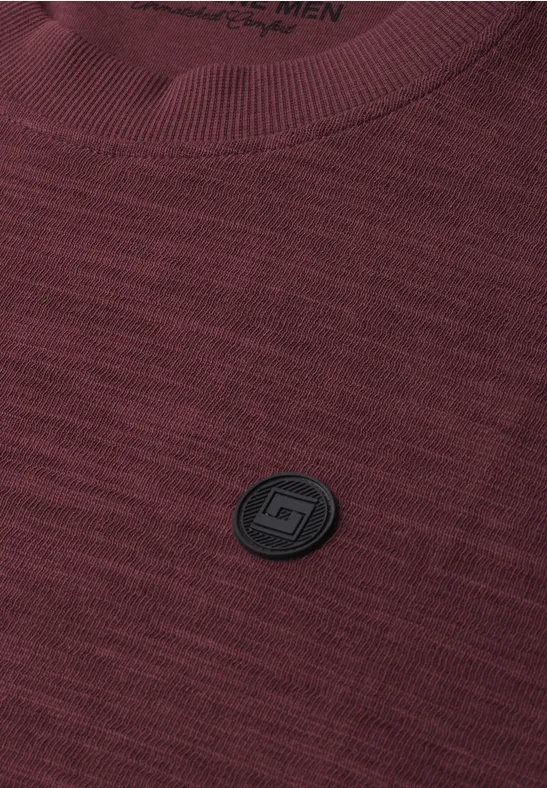 Bordeauxfarbenes gestricktes T-Shirt mit schwarzem rundem Logoemblem auf der Brust.