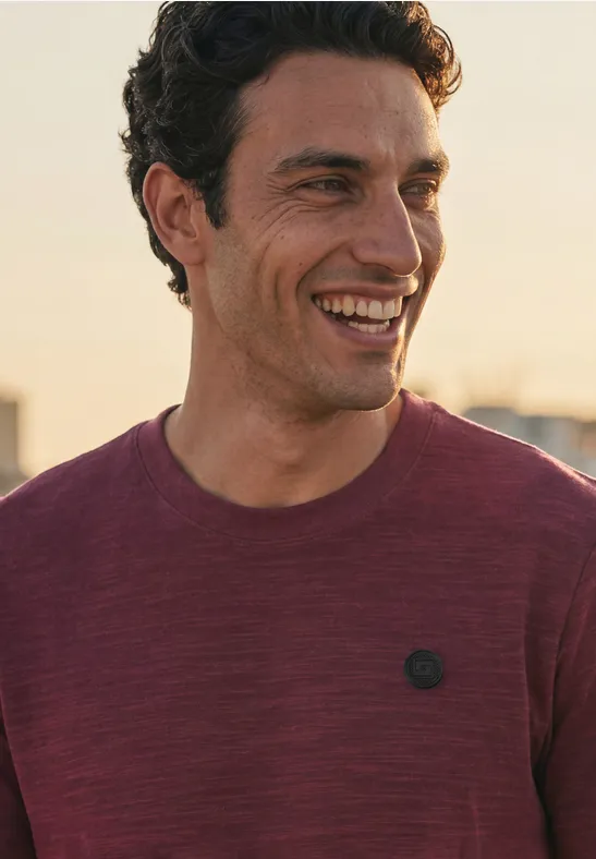 Ein lächelnder Mann trägt ein bordeauxrotes T-Shirt mit kleinem Logo.