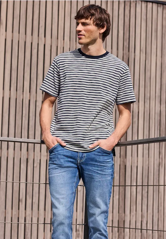 Ein Mann steht entspannt vor einem Holzzaun, trägt gestreiftes T-Shirt und Jeans.