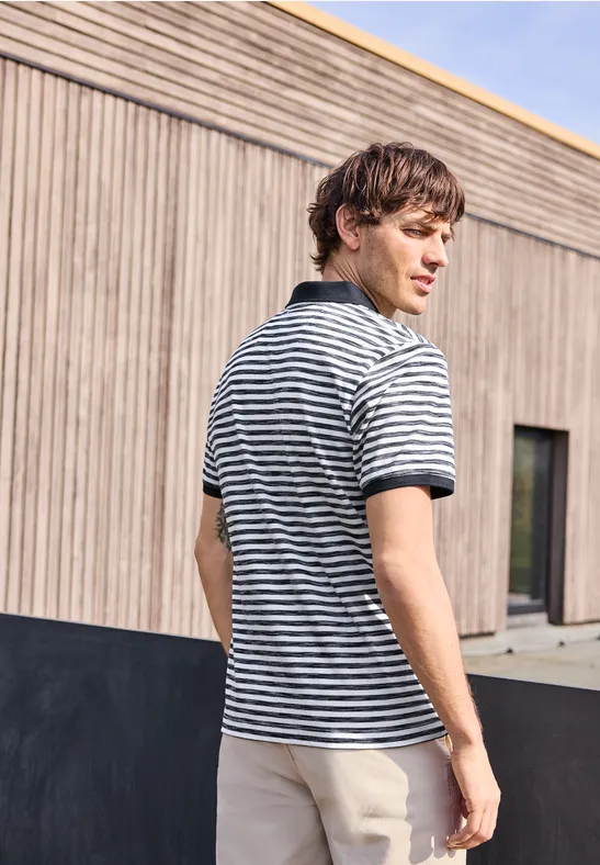 Ein junger Mann in gestreiftem Poloshirt und beigen Hosen steht mit abgewandtem Blick vor einer Holzwand.