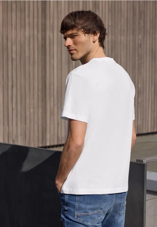 Een jonge man staand in profiel voor een houten wand, met een wit T-shirt en een blauwe spijkerbroek.