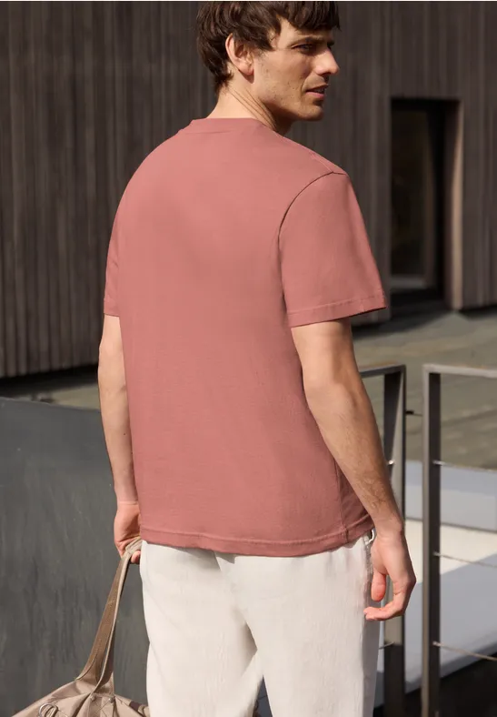 Een man van achteren in een roze T-shirt en lichte broek, met een tas voor een moderne houten gevel.