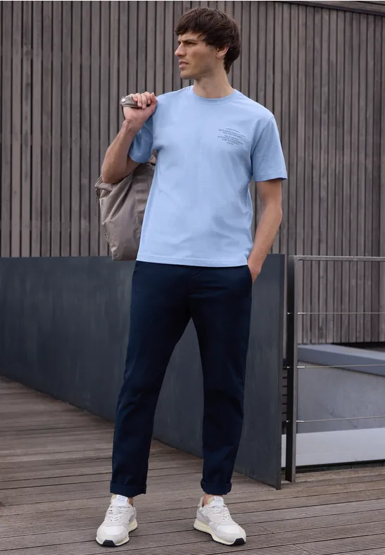 Ein Mann trägt lässiges Outfit mit hellblauem T-Shirt, dunkelblauen Hosen und weißen Sneakern draußen.