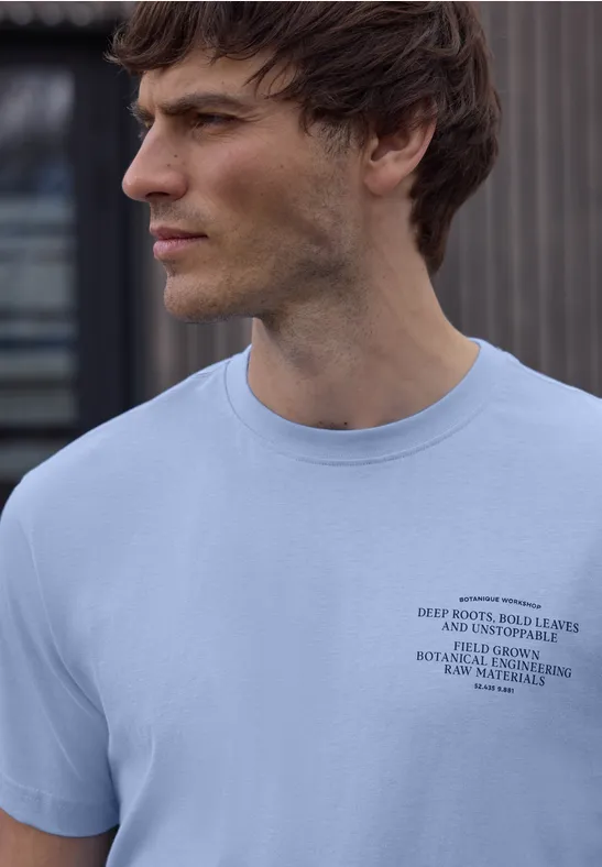 Ein seitlich abgebildeter Mann trägt ein hellblaues T-Shirt mit kleinem schwarzen Logo, unscharfes Gesicht.