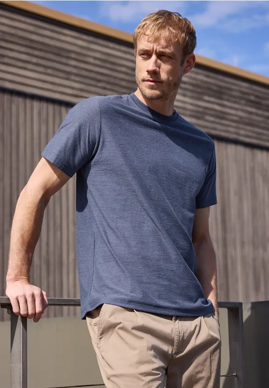 Mann lehnt an Geländer vor Holzwand in blauem T-Shirt