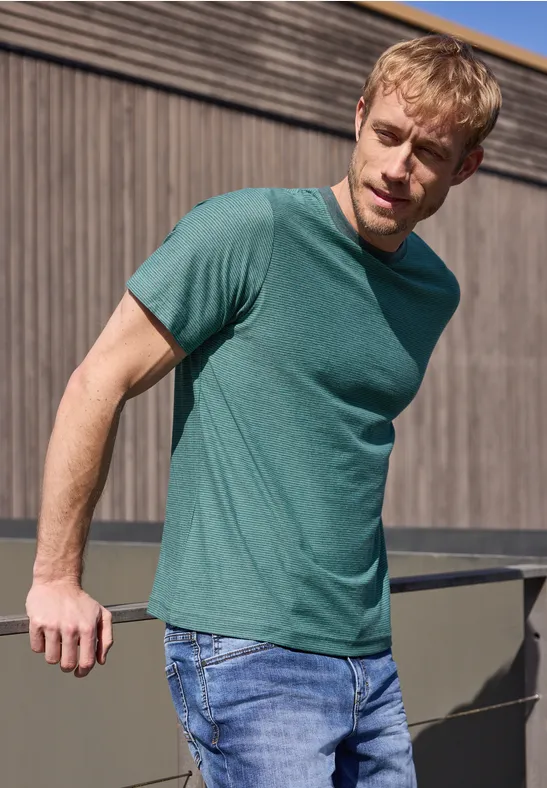 Ein Mann in einem grünen T-Shirt und Jeans lehnt lässig an einer Brüstung vor einer Holzwand.