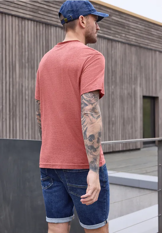 Ein Mann von hinten in rotem T-Shirt, Jeans-Shorts, Baseballcap und auffälligen Tattoos auf dem Arm.
