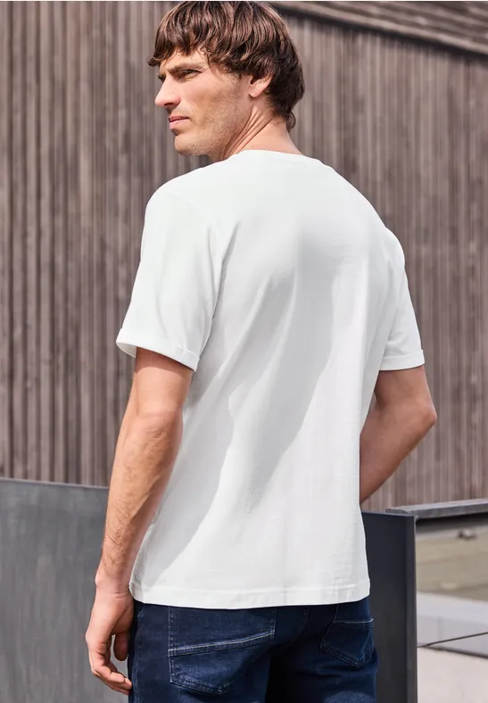 Ein Mann steht seitlich gesehen, trägt ein weißes T-Shirt und blaue Jeans vor einer Holzfassade.