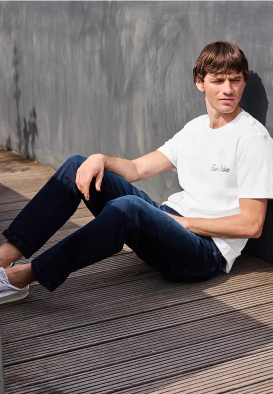 Ein junger Mann sitzt lässig in weißem T-Shirt und Jeans auf einem Holzdeck vor einer grauen Wand.