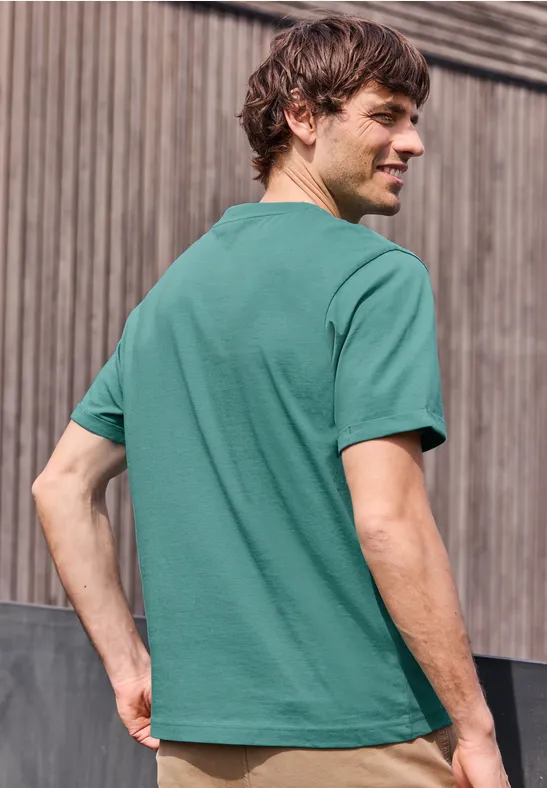 Ein Mann steht mit dem Rücken zur Kamera, trägt ein grünes T-Shirt vor einem Holzzaun.