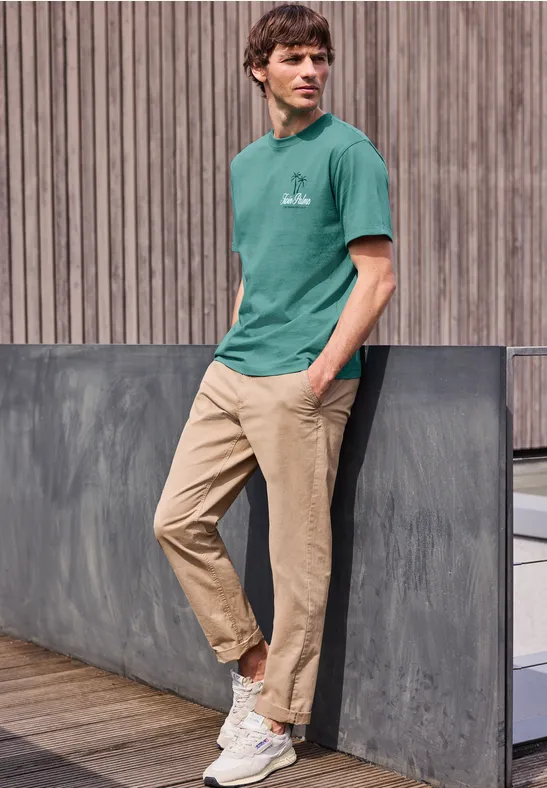 Junger Mann in grünem T-Shirt und beigen Chinos lehnt entspannt an einer Betonmauer vor Holzfassade.