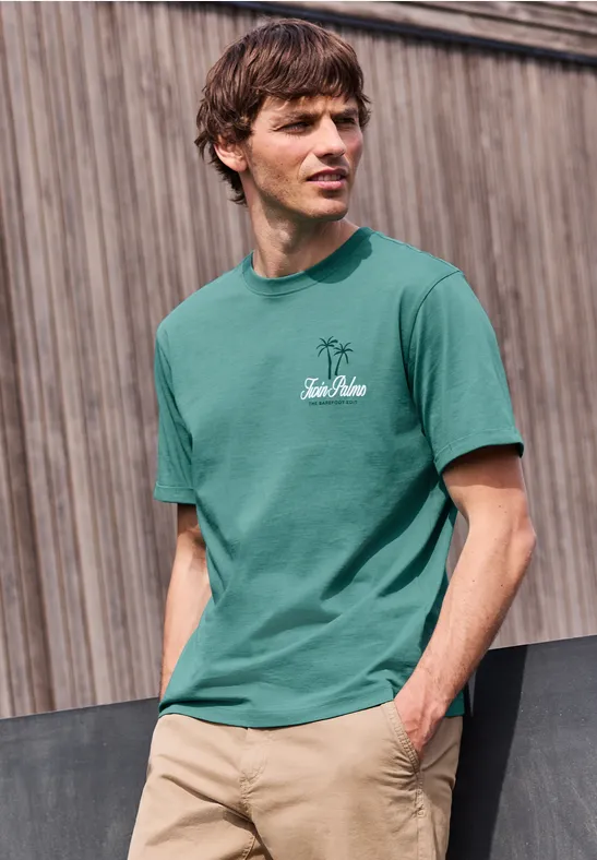 Junger Mann in grünem T-Shirt und beiger Hose lehnt vor einer Holzfassade.