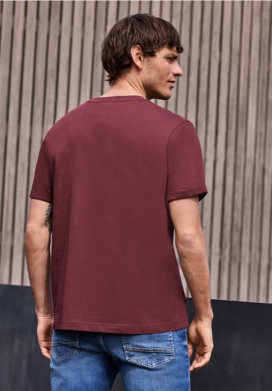 Ein Mann steht mit dem Rücken zur Kamera, trägt ein weinrotes T-Shirt und Jeans vor einer Holzlattenwand.