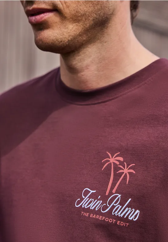 Nahaufnahme eines Mannes in bordeauxrotem T-Shirt mit Palmenmotiv und Schriftzug „Twin Palms“