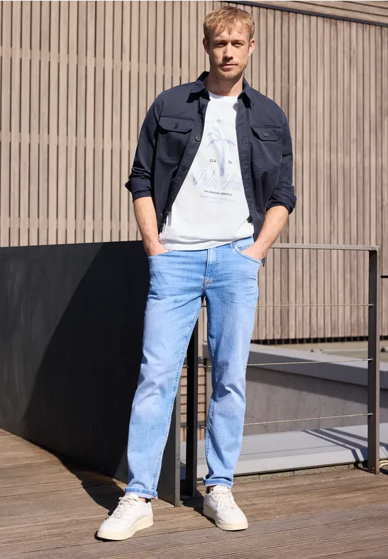 Een jonge man leunt ontspannen tegen een terrasbalustrade, gekleed in jeans en sneakers.