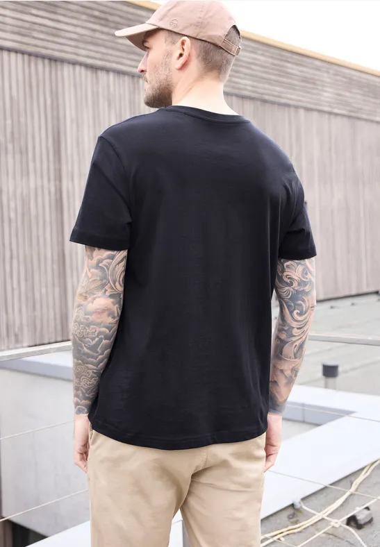 Mann von hinten auf Dach, schwarzes T-Shirt, beige Hose, tätowierte Arme, rosa Mütze.