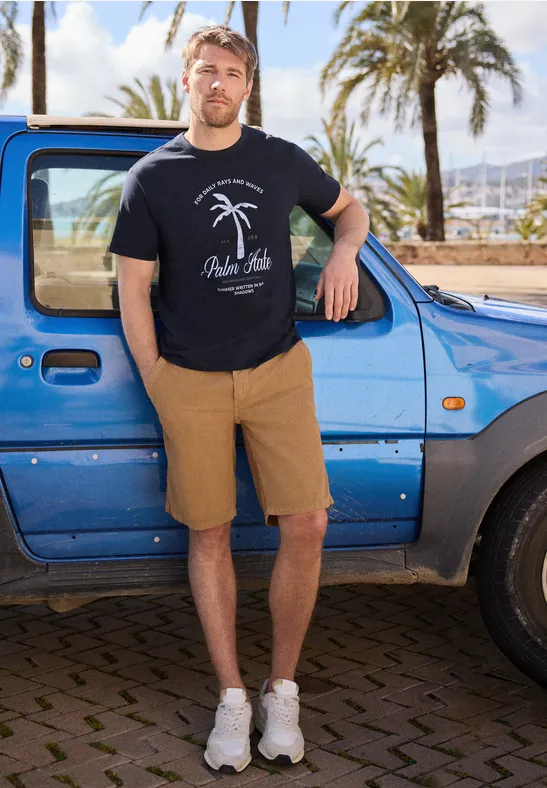 Ein Mann lehnt in Shorts und T-Shirt an einem blauen Auto, im Hintergrund Palmen und sonnige Atmosphäre.