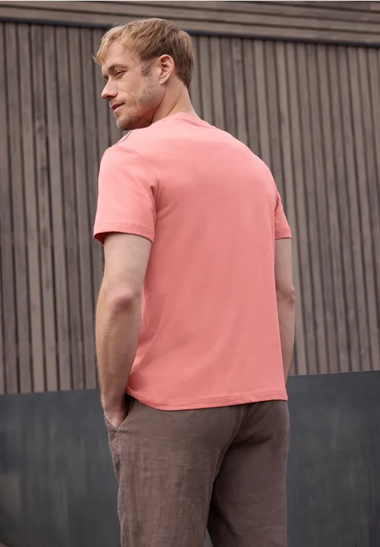 Man in zalmroze T-shirt en bruine broek zijwaarts tegen een houten muur geleund.