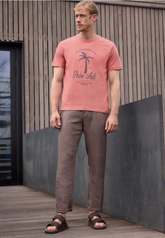 Man in roze T-shirt met palmprint, bruine broek en sandalen op een houten terras.