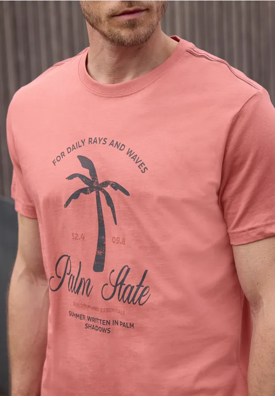 Een man draagt een zalmkleurig T-shirt met een palmbomenprint en de tekst “Palm State”.