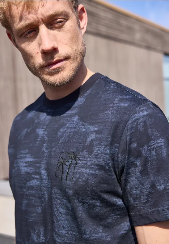 Man in donkerblauw T-shirt met palmborduring, staand voor een houten muur buiten.