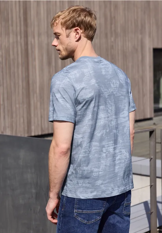 Een man van achteren met een grijs-blauw T-shirt staat voor een houten wand buiten.