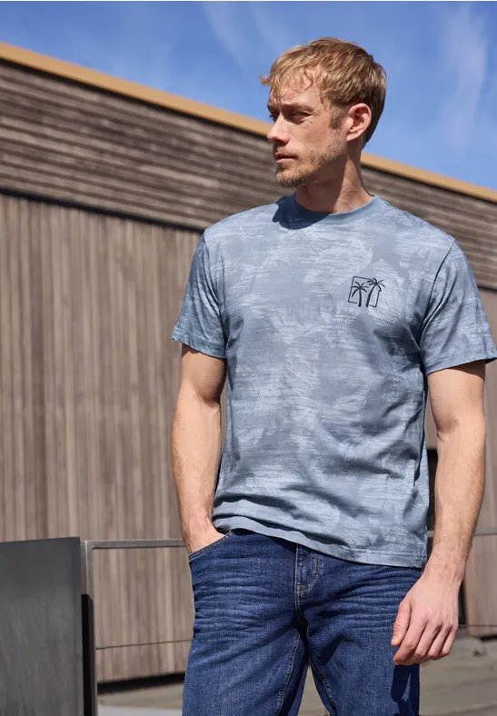 Een man staat voor een houten wand, draagt een grijs tie-dye t-shirt en spijkerbroek, handen in de zakken.