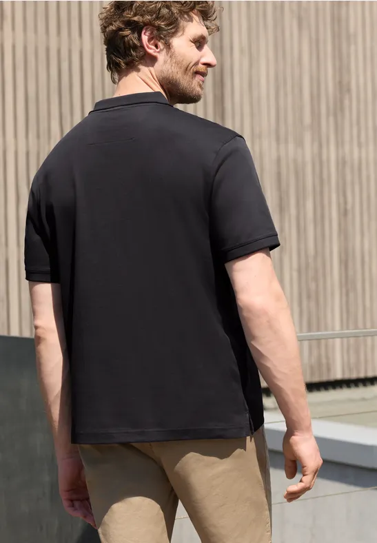 Een man van achteren draagt een zwarte polo en een beige korte broek voor een houten gevel.