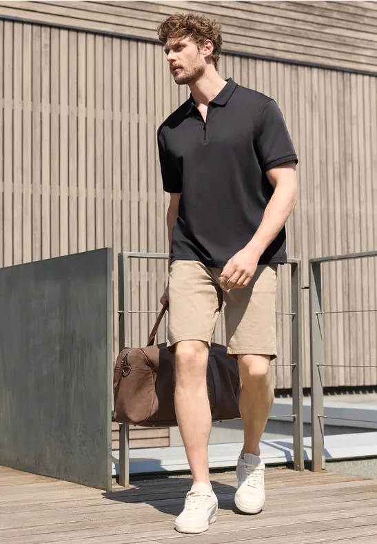 Een man in een zwart poloshirt, kaki short en witte sneakers draagt een reistas terwijl hij langs een modern gebouw loopt.