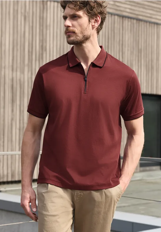 Een man draagt een bordeauxrood poloshirt met korte mouwen en rits, en een lichte broek voor een houten gevel.
