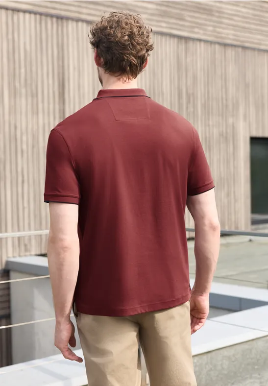 Een man van achteren in een bordeaux polo en lichte broek staat voor een houten gevel.