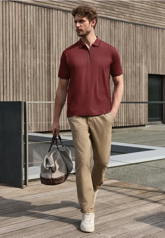 Man in bordeaux poloshirt, beige broek en witte sneakers loopt met reistas langs een modern gebouw.
