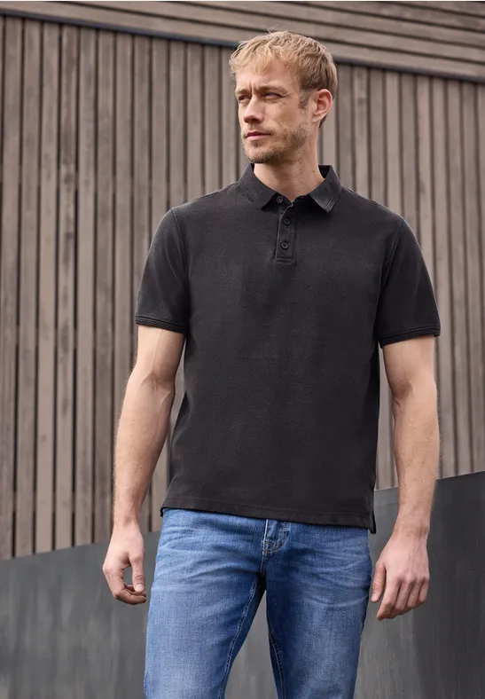Ein Mann in schwarzem Poloshirt und blauen Jeans steht vor einer Holzfassade.