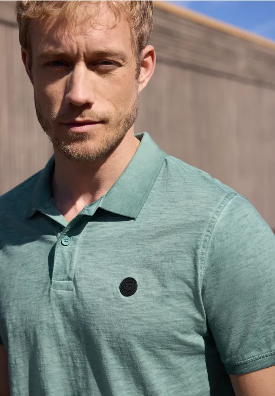 Ein Mann in grünem Poloshirt steht vor einer Holzwand, sein Gesicht ist unkenntlich gemacht.